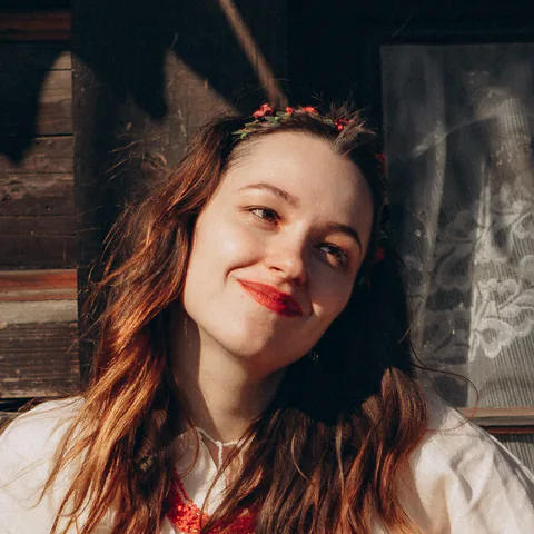 Anna Kulak in a white embroidered blouse and a floral headband, by the window of an old wooden house